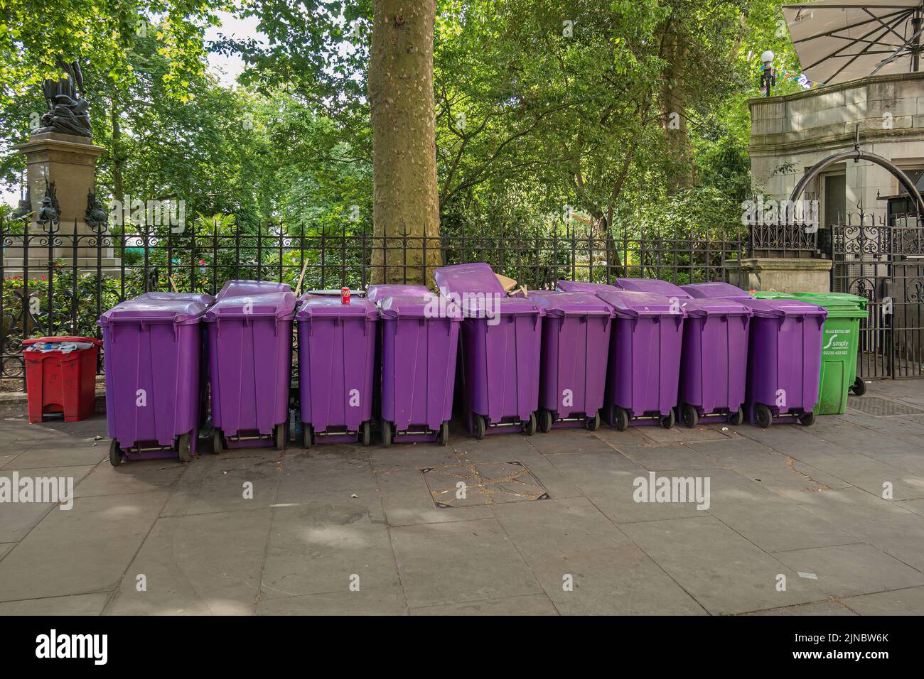 London, UK- July 4, 2022: Row of purple garbage cans outside Whitehall gardens. Green foliage and Sir James Outram statue in back. Stock Photo