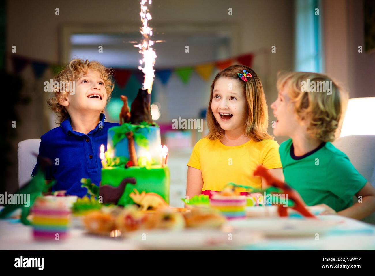 Kids birthday party. Dinosaur theme cake. Little girl blowing candles and opening gifts. Children event. Decoration for dinosaurs themed celebration. Stock Photo