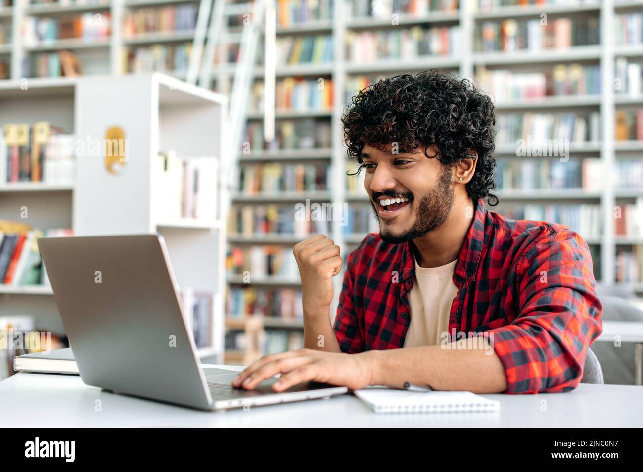 Cheerful amazed indian or arabian guy, student, sitting in university library with laptop, rejoices in success, win, good mark on the exam, gesturing hand, smiling, emotional face expression Stock Photo