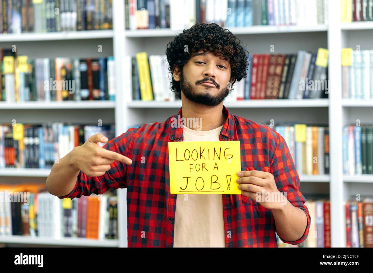 Upset curly indian or arabian unemployed guy, stands in the library against the background of bookshelves, holds a sign in his hand that says looking for a job, looks at camera, hopes to get a job Stock Photo