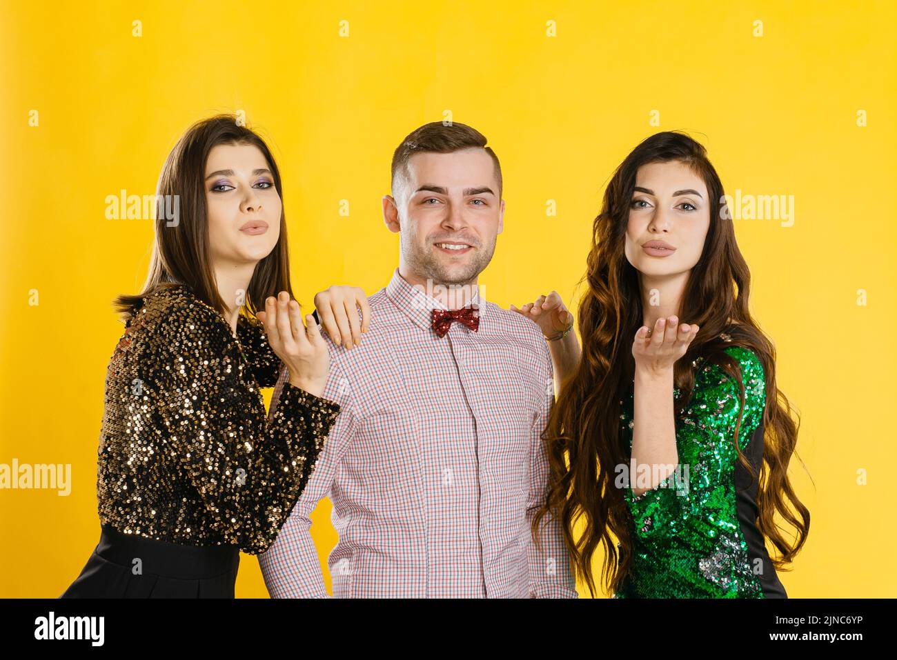 Young adult girls hug a guy and make an air kiss to the camera. Festive corporate party of friends. A man with two girls on a yellow background in fes Stock Photo