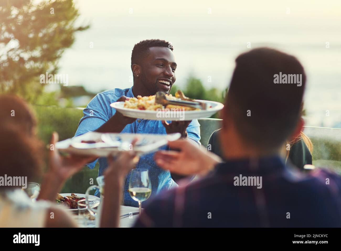 Heres some more for you mate. a handsome young man passing a plate of pasta around at a gathering with friends outdoors. Stock Photo