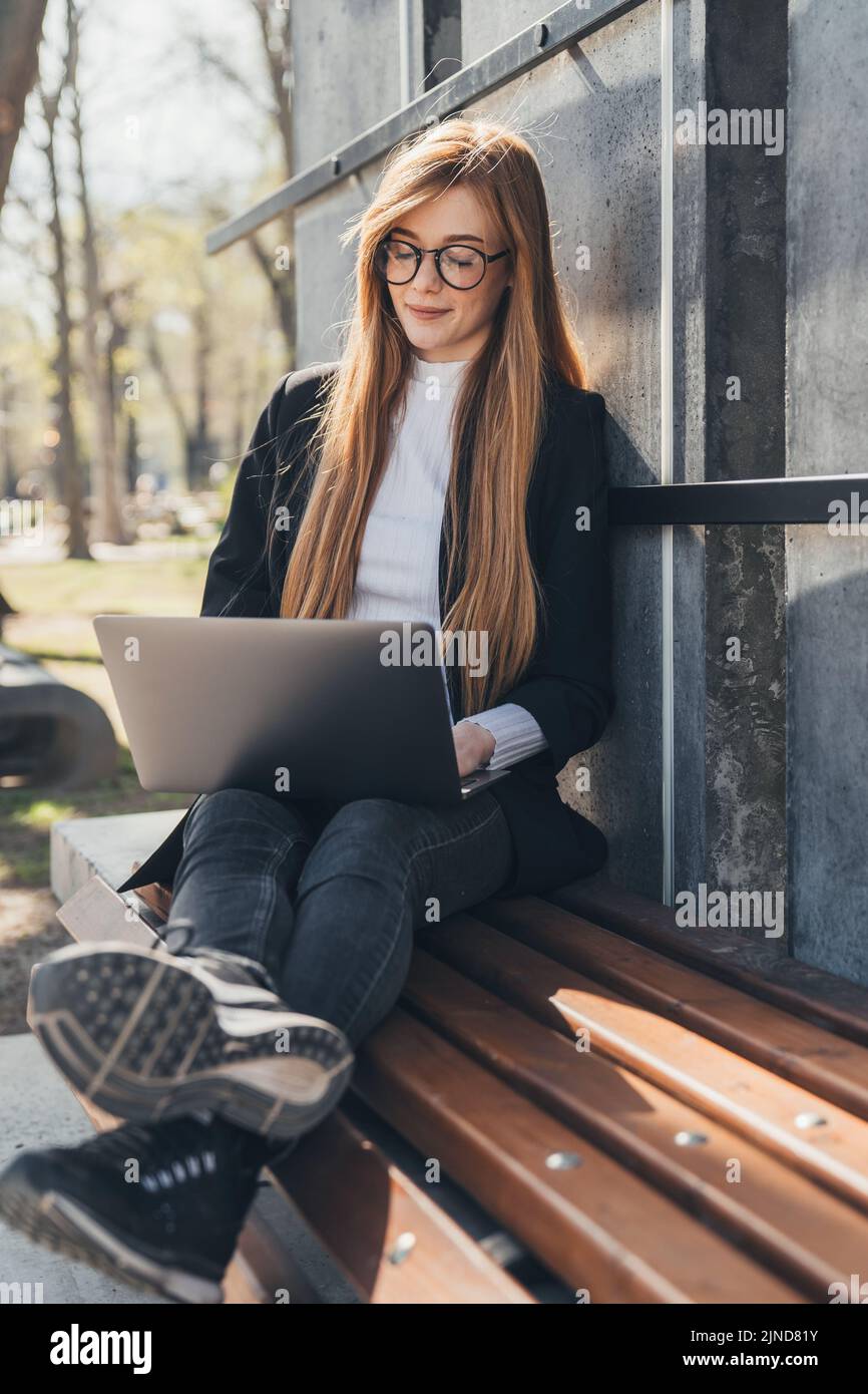 Caucasian professional businesswoman sitting on a bench with computer laptop working outside in the city park. Stock Photo