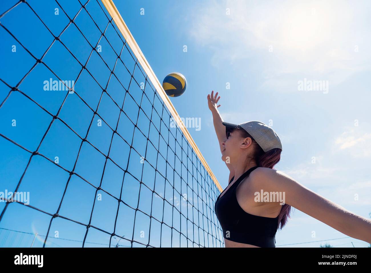 Girl athlete jumping in front of the yellow net for the ball while playing volleyball. Blocking the ball. Beach volleyball in the open air. Stock Photo