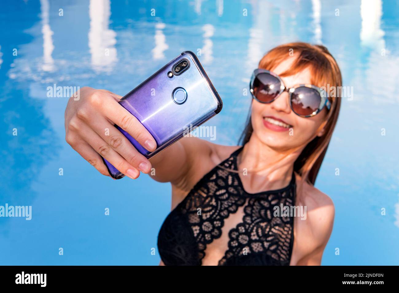 Young beautiful woman taking pictures of herself on a mobile phone on the background of clear clear water in the pool. Stock Photo