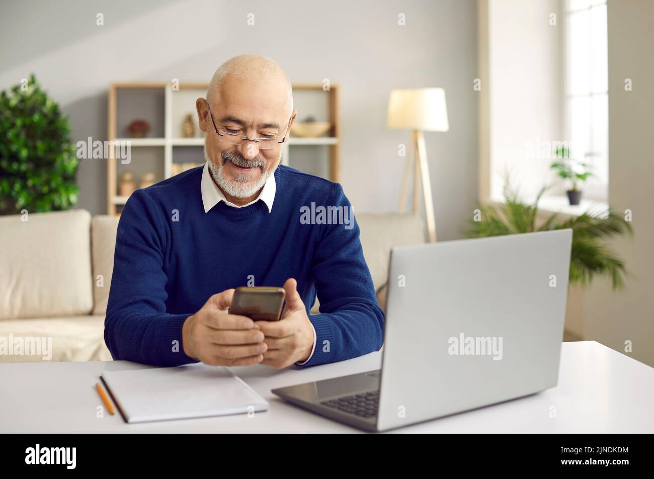 Happy senior man using his mobile phone while sitting at desk with laptop computer Stock Photo