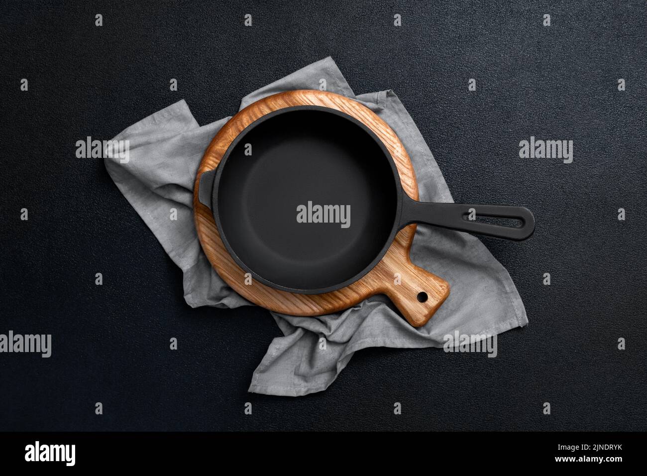 Cast iron skillet on a wooden board. Frying pan, cutting board and kitchen towel on a black background. Kitchen utensils. Empty place. Top view. Stock Photo