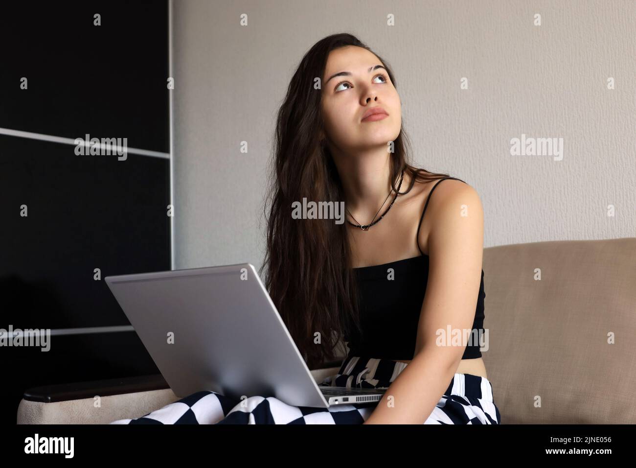 Pretty young girl with long hair wearing black top sitting with laptop on sofa and looking up. Concept of inspiration during work, study or leisure Stock Photo