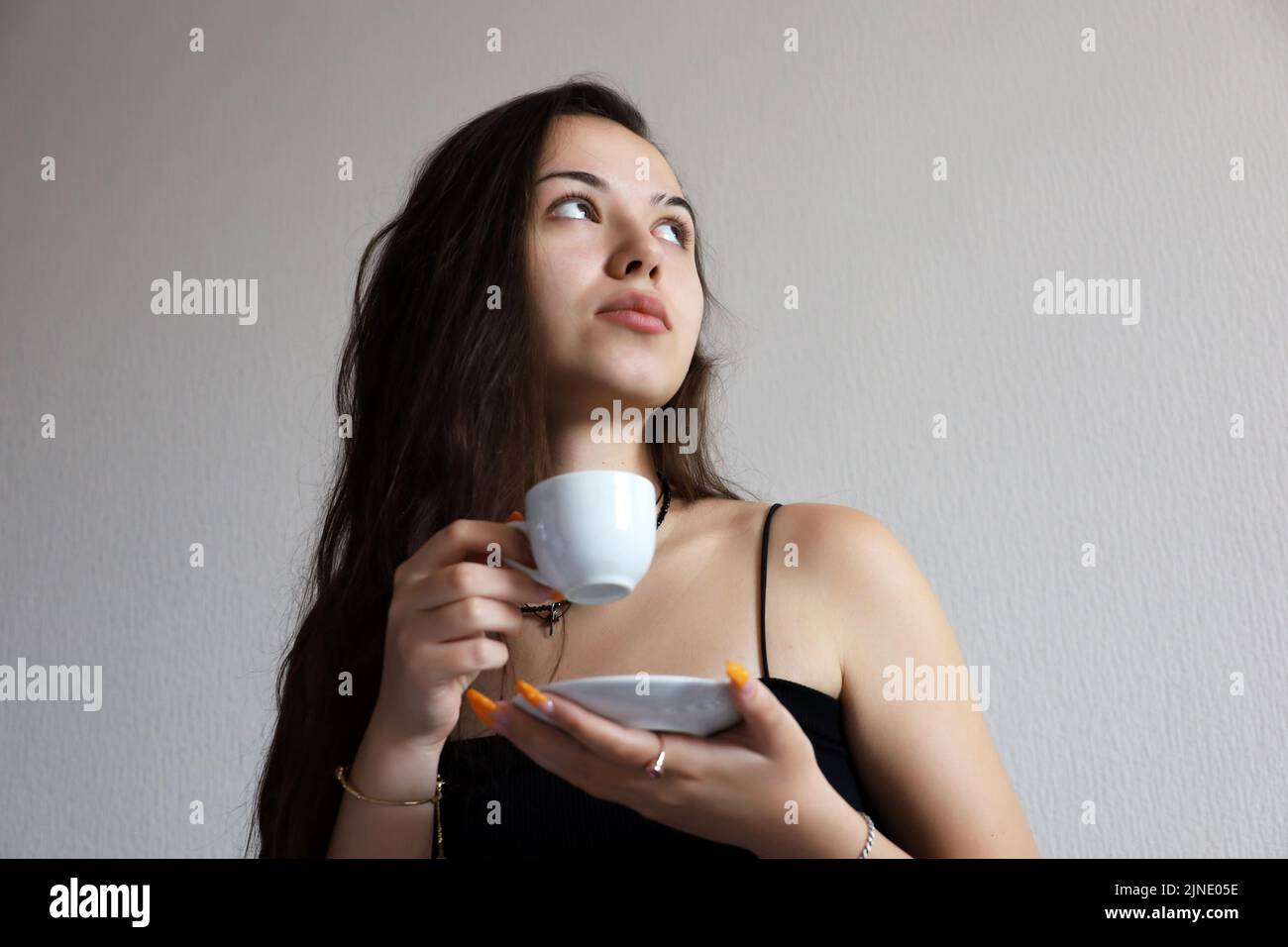Sensual young girl with long hair drinking tea or coffee, white cup and saucer in female hands. Concept of inspiration, relaxing at home, cozy morning Stock Photo