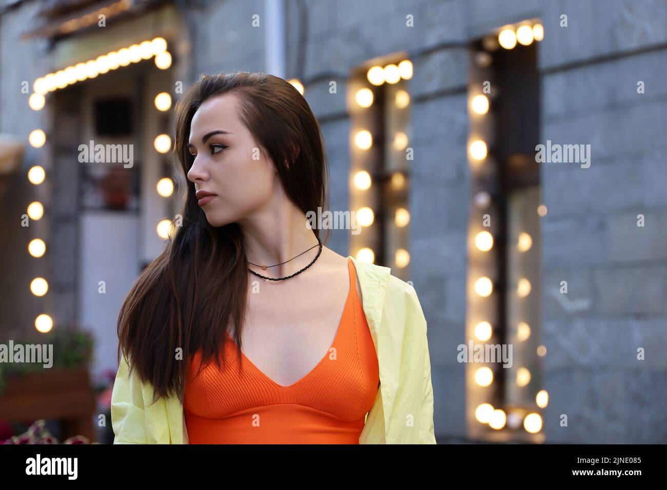 Portrait of pretty young girl with long hair and perfect makeup wearing orange top and yellow shirt on a street on blurred lights background Stock Photo