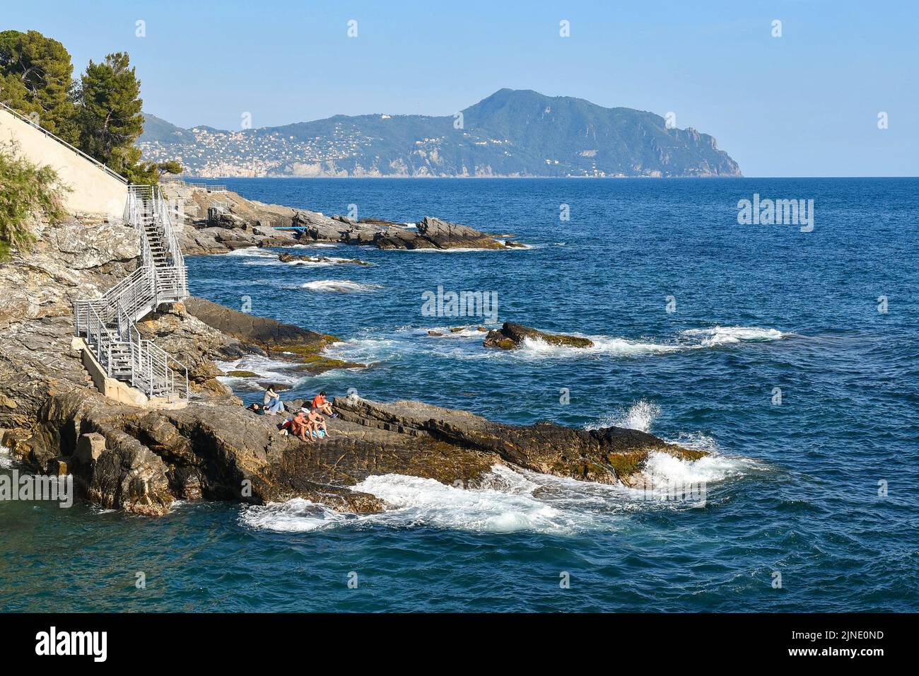 People sunbathing on the rocks by the sea of the Anita Garibaldi Promenade with the promontory of Portofino in the background, Nervi, Genoa, Liguria Stock Photo