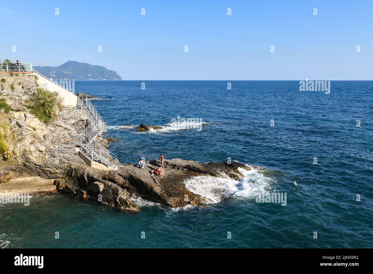 People sunbathing on the rocks by the sea of the Anita Garibaldi Promenade with the promontory of Portofino in the background, Nervi, Genoa, Liguria Stock Photo