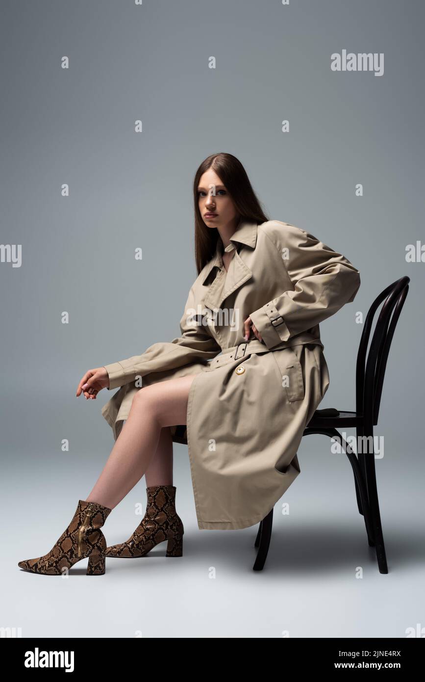full length of brunette model in trench coat sitting on chair and ...