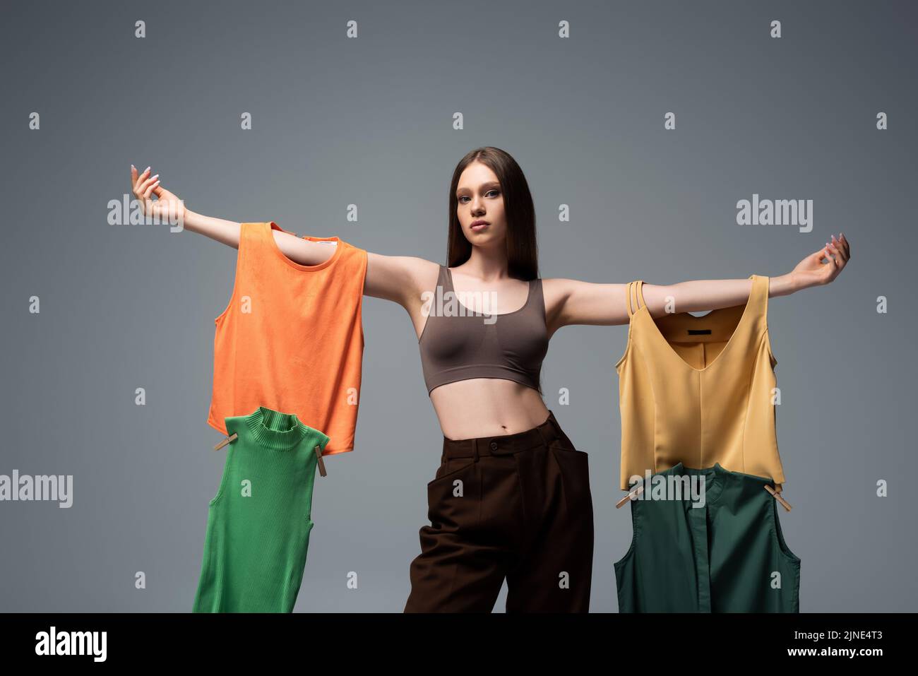 pretty model in crop top and trousers standing with outstretched hands and holding clothing on grey Stock Photo