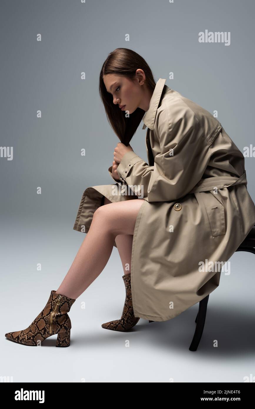 full length of model in stylish trench coat sitting on chair and posing ...