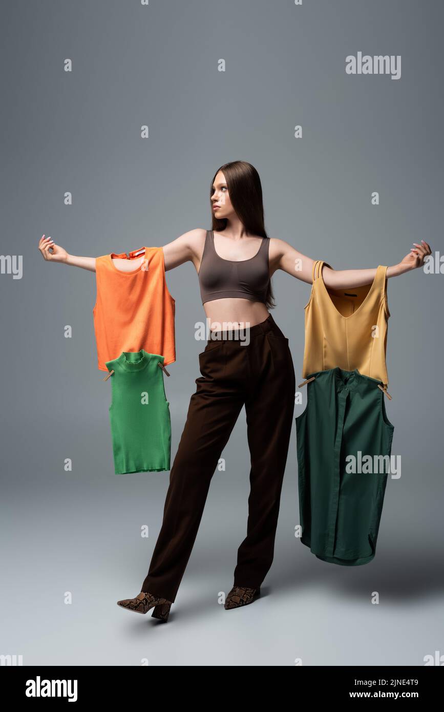full length of pretty model in crop top and trousers standing with outstretched hands while holding clothing on grey Stock Photo