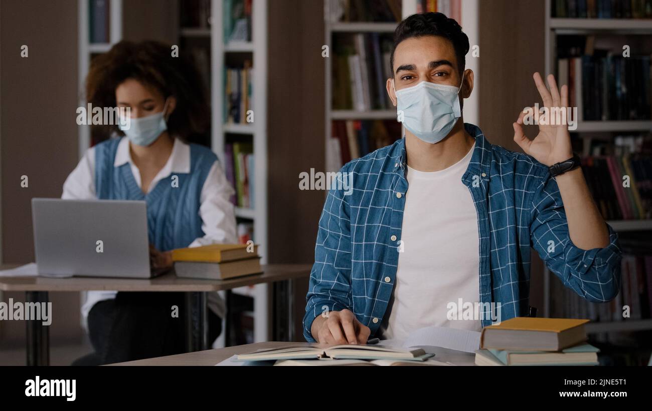 Young clever guy student in protective mask writes notes prepares for exam makes project writing essay shows ok sign approval gesture advises higher Stock Photo