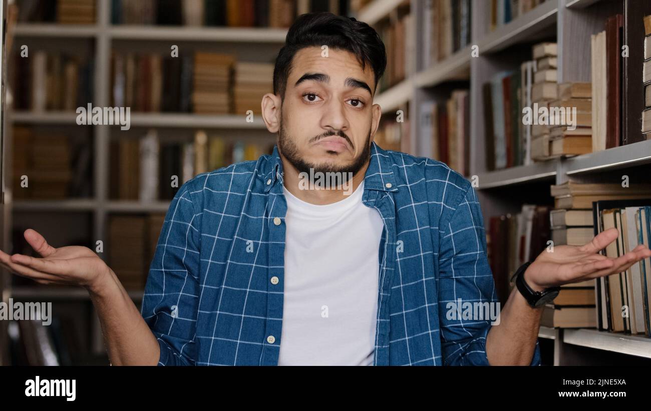 Close-up young upset man standing indoors in university library hispanic student misunderstood puzzled shrugs shoulders doesnt know what to answer dou Stock Photo