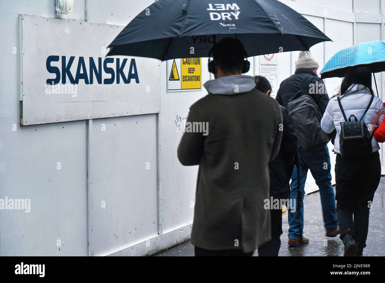 London, United Kingdom - February 01, 2019: Pedestrians with umbrellas walking near wooden barrier at construction site with label Skanska. It is Swed Stock Photo