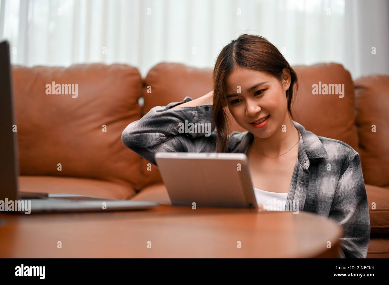 Pretty Asian young woman uses digital tablet touchpad to chat with her online friends while relaxing in her living room. Stock Photo