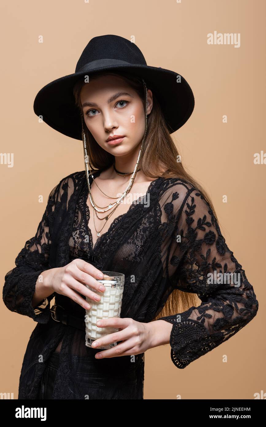 Stylish woman in fedora hat holding glass of milk isolated on beige