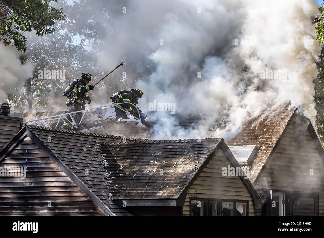 Two  Sag Harbor firefighters operating off of an aerial ladder use a chain saw and a hook to cut a hole in the roof of the building as Sag Harbor Fire Stock Photo