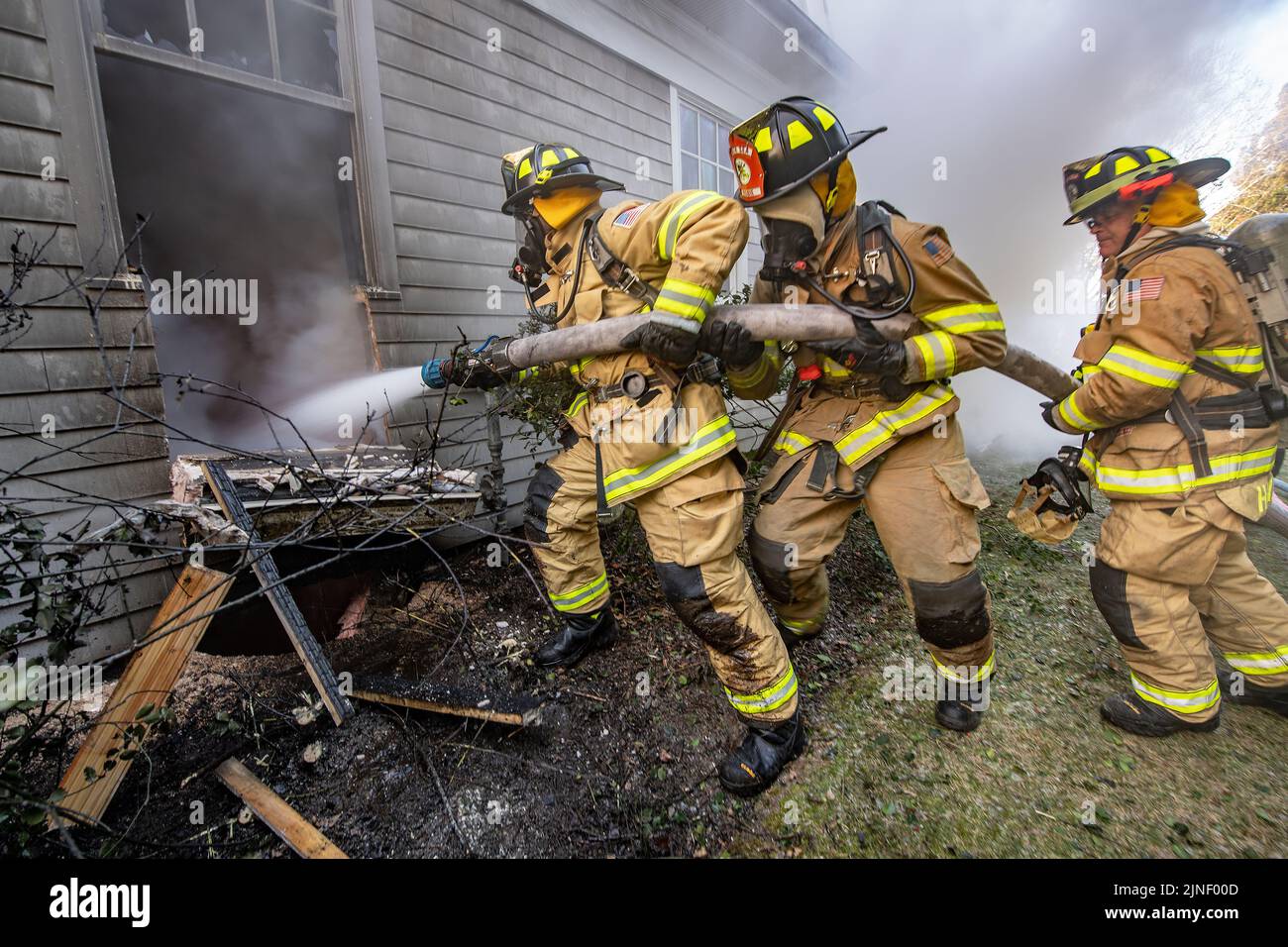 A three-man hose team uses a hose to spray water through a window to try to extinguish the fire within it as the Bridgehampton Fire Department, with m Stock Photo