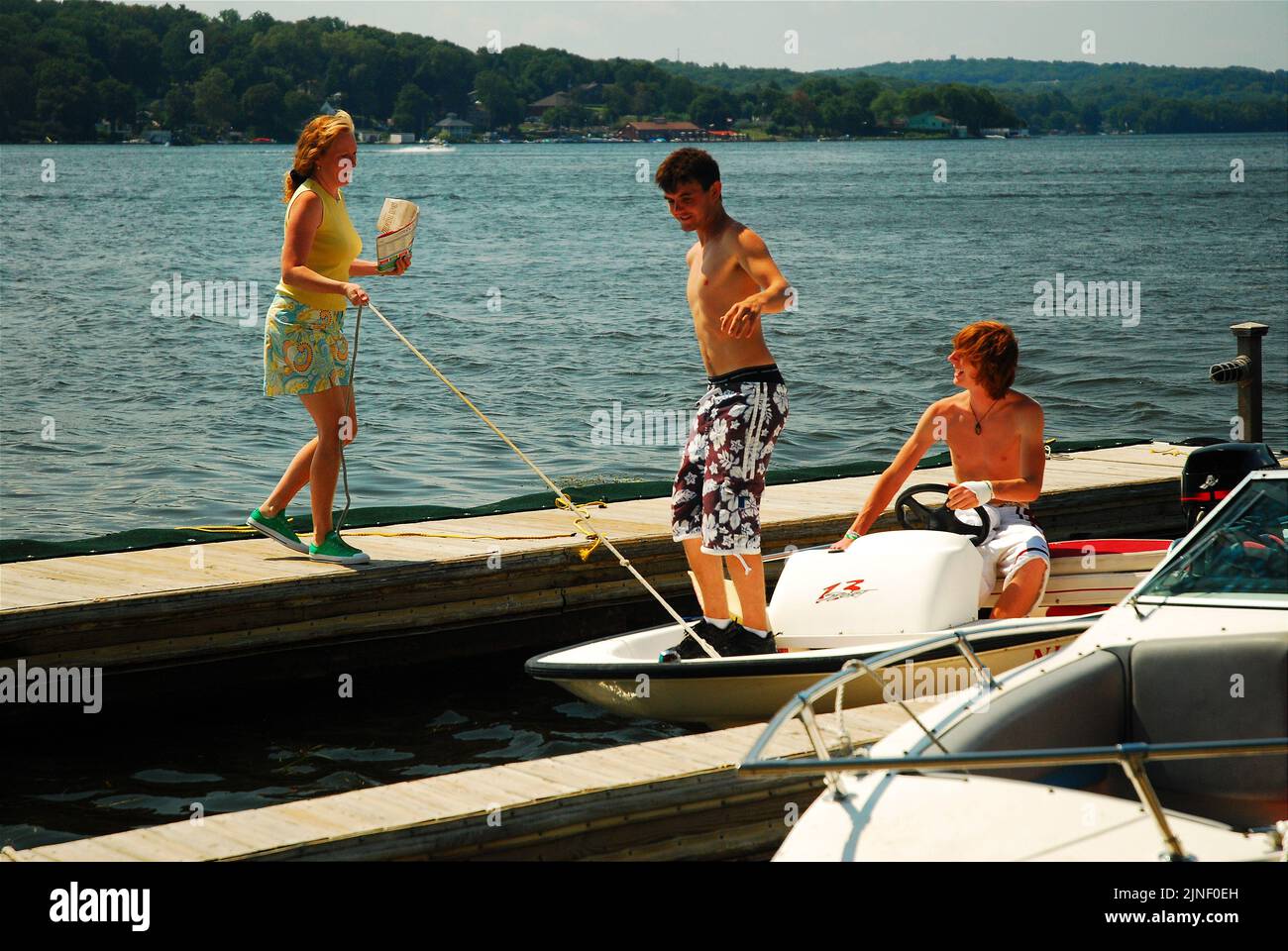 An adult woman helps two teenage boys bring their small boat into the dock for tying on a beautiful summer vacation day on  the lake Stock Photo