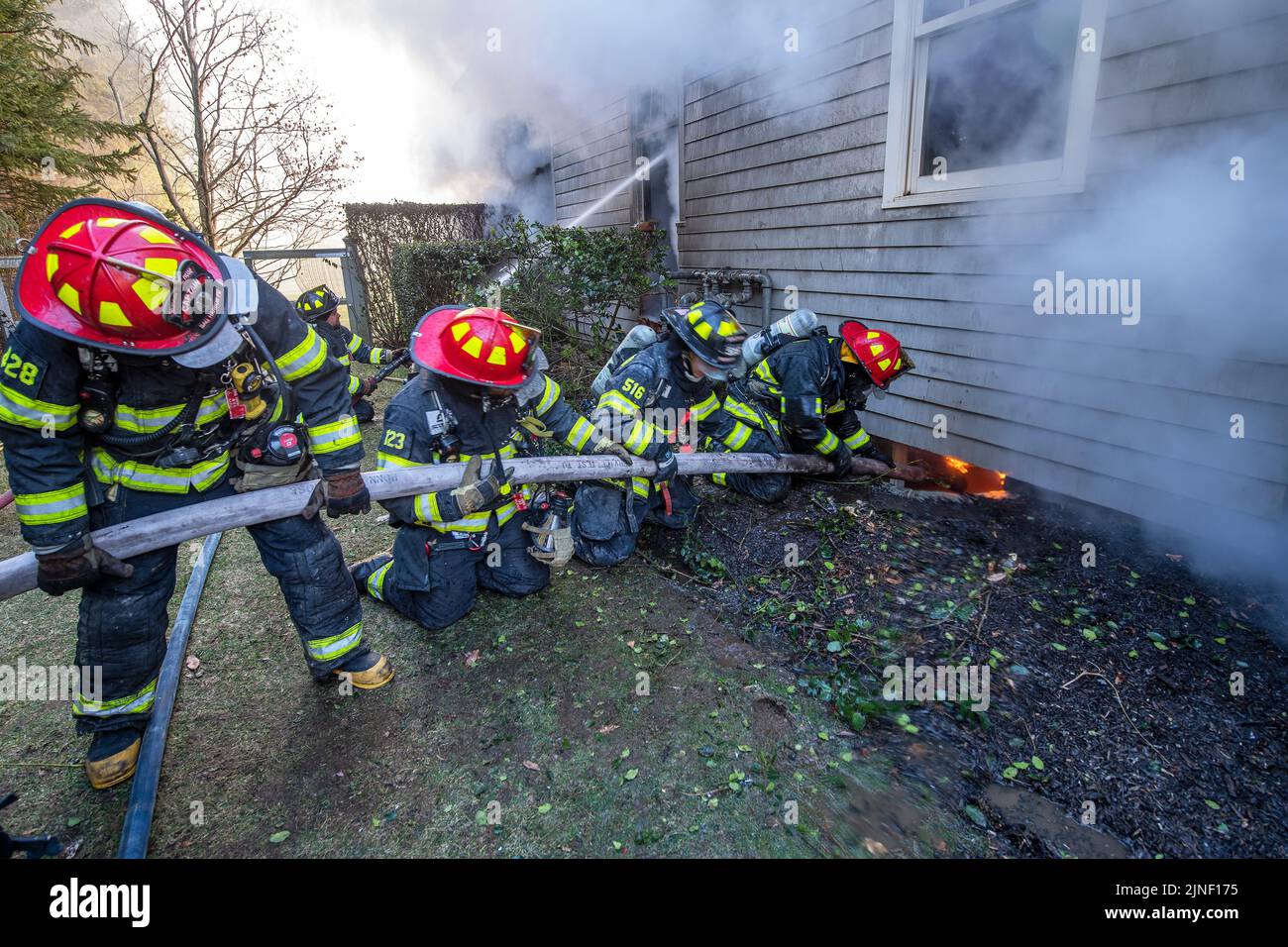 A four-man hose team uses a hose to spray water through a basement window to try to extinguish the fire within it as the Bridgehampton Fire Department Stock Photo