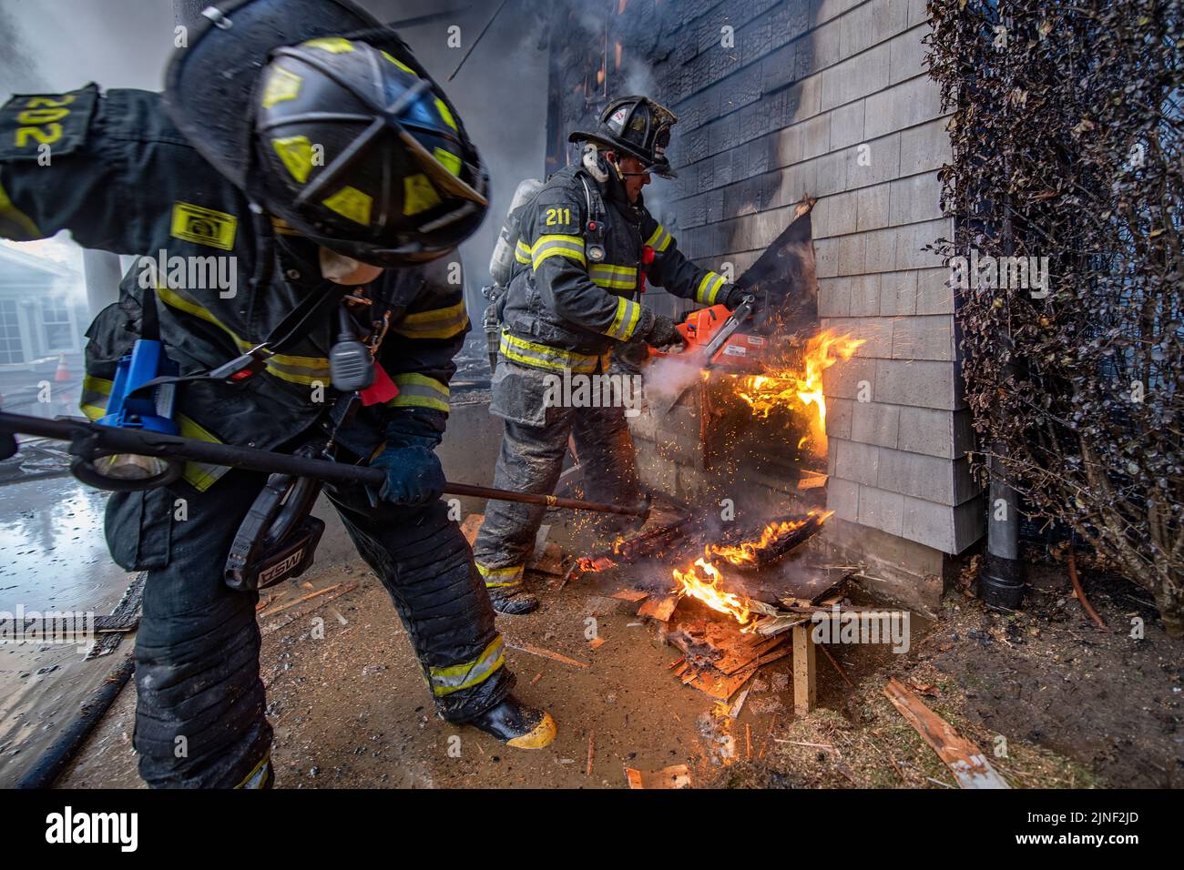 Sag Harbor firefighters use a chain saw, and a hook to open up an exterior wall to expose the fire within it as the Bridgehampton Fire Department, wit Stock Photo