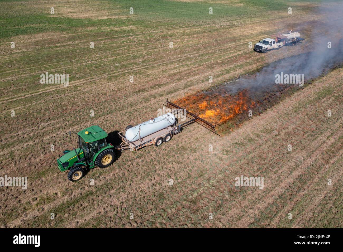 A tractor pulls a propane burner to burn weeds in an hay field after cutting the alfalfa on a ranch in Utah.  A water truck follows to control the fir Stock Photo