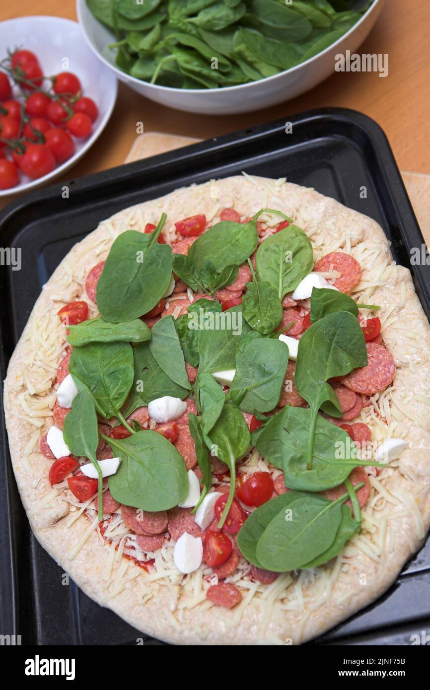 Fresh pizza ready to go into the oven - shallow dof with bowls of tomatoes & spinach in the background Stock Photo