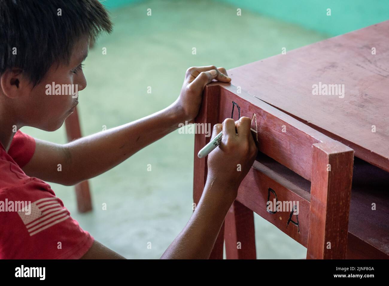 A riberenos student carves his initials into a donated desk and chair in the Peruvian Amazon Stock Photo