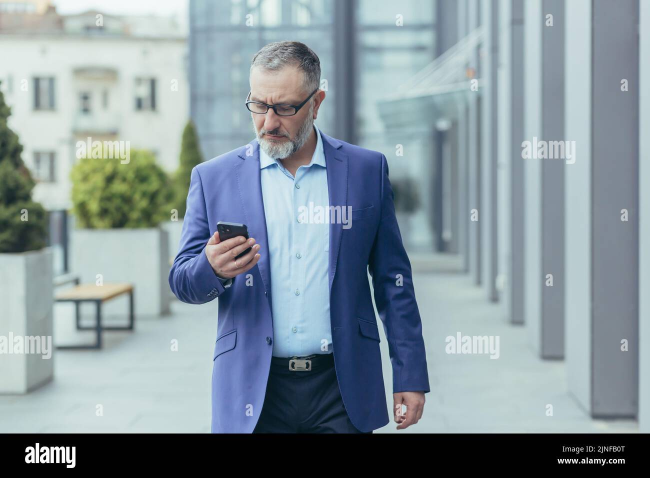 Serious and successful senior gray-haired businessman walking outside office building, man holding phone, thoughtfully reading news Stock Photo