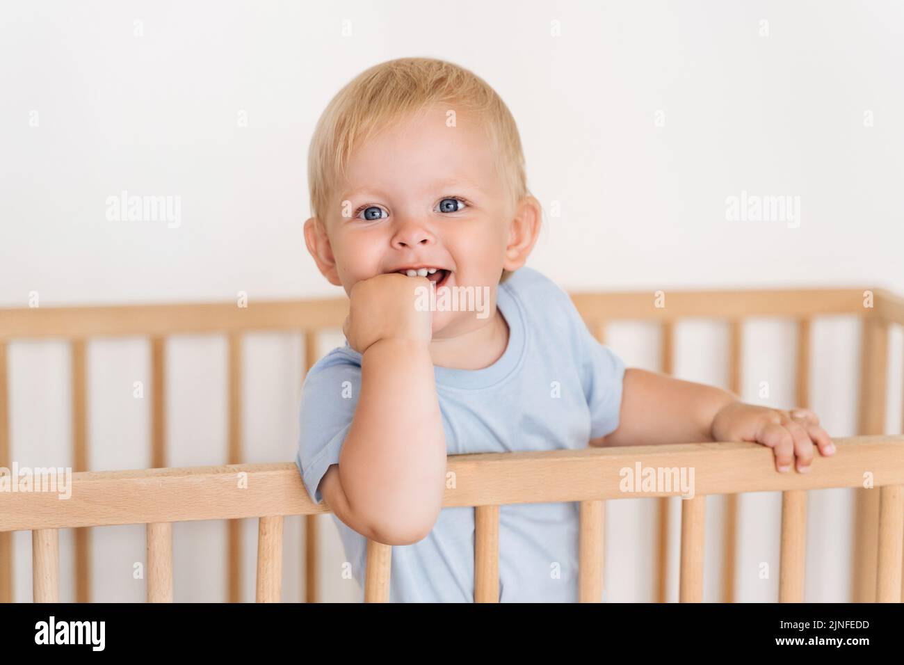 Indoor portrait of happy blond baby boy on white background growing first milk tooth standing in wooden bed with hand in mouth massaging itchy gums. Teething concept Stock Photo