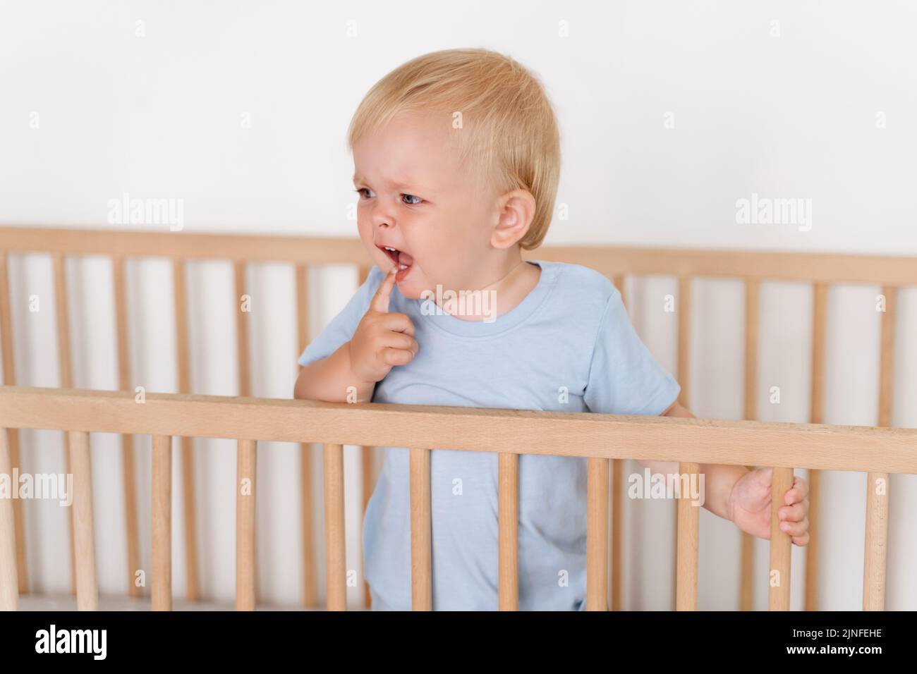 Portrait of cute baby boy standing in bed crying while touching itchy gums, growing new milk tooth suffering from teething pain. Childhood and childcare concept. Health care Stock Photo