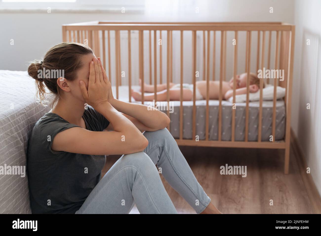 Side view of mother covering face crying from exhaustion sitting on floor next to baby crib while child napping, can't handle difficulties of maternity, having postnatal depression symptoms Stock Photo