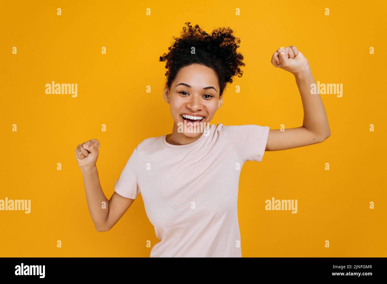 Joyful happy curly-haired african american woman, dancing, gesturing with fists, receives profit, glad to win lottery, standing on isolated orange background. Good news, success and happiness concept Stock Photo