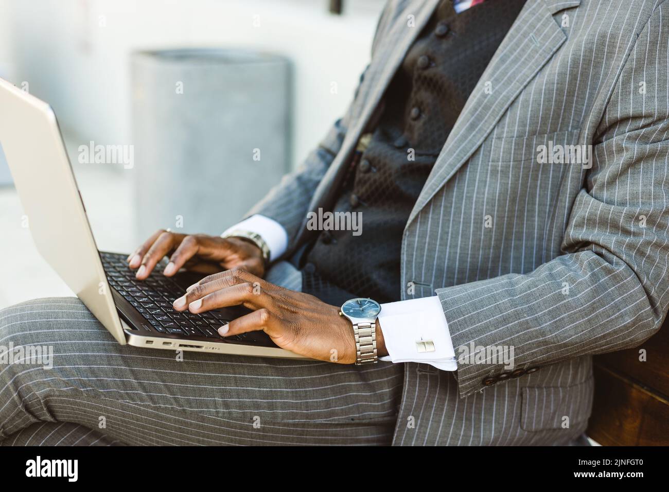 A black businessman in a suit uses a laptop outdoors while sitting on a bench. Close-up Stock Photo