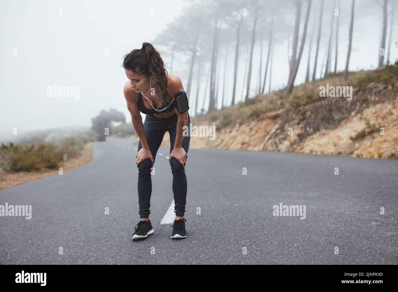 Athletic young woman taking a break from jogging outdoors. Sporty young woman working out along a misty road in the morning. Stock Photo