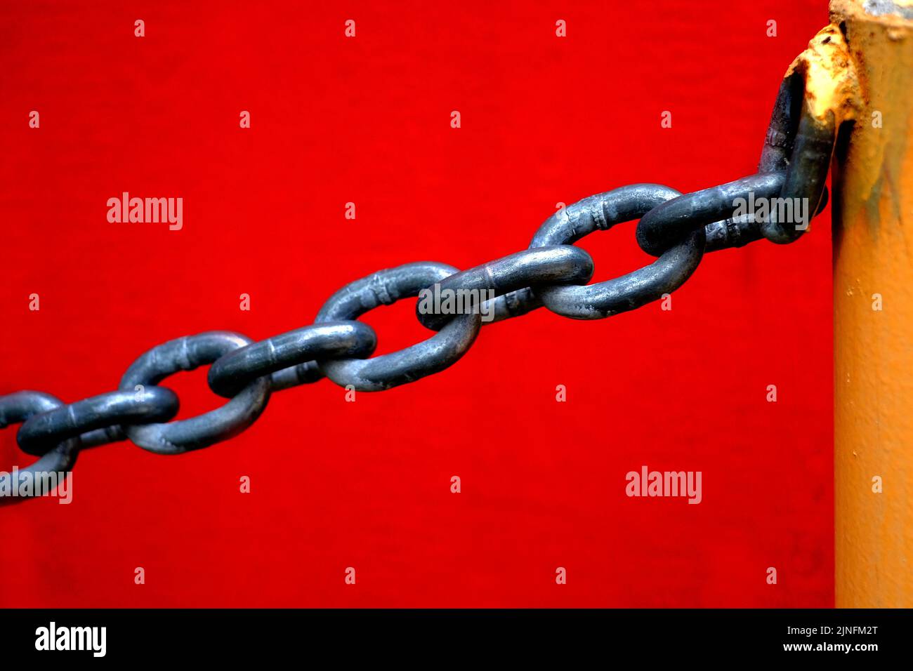 Chain for security hanging from yellow metal pole with red wall in background Stock Photo