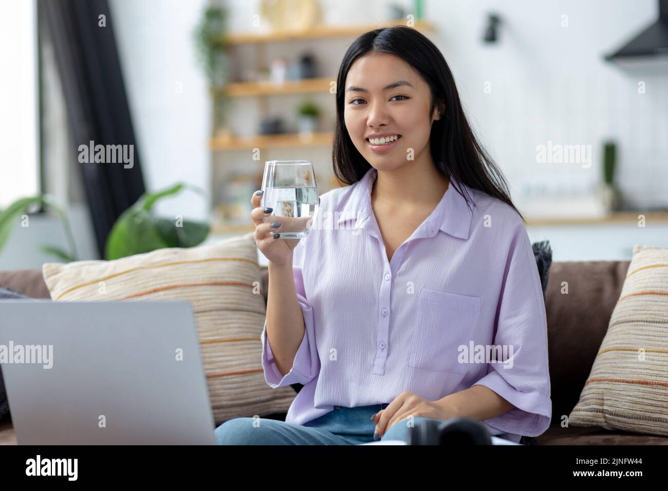 female freelancer with glass fresh water sitting at sofa Smiling woman using laptop computer working at home Stock Photo