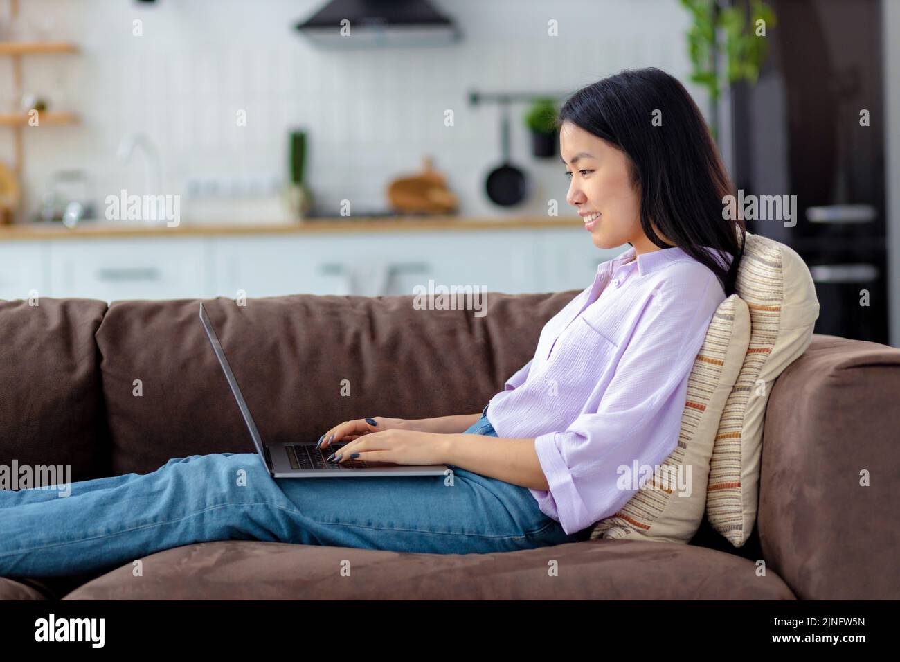 Happy Asian woman using laptop on sitting of cozy sofa at home chatting on social network Stock Photo
