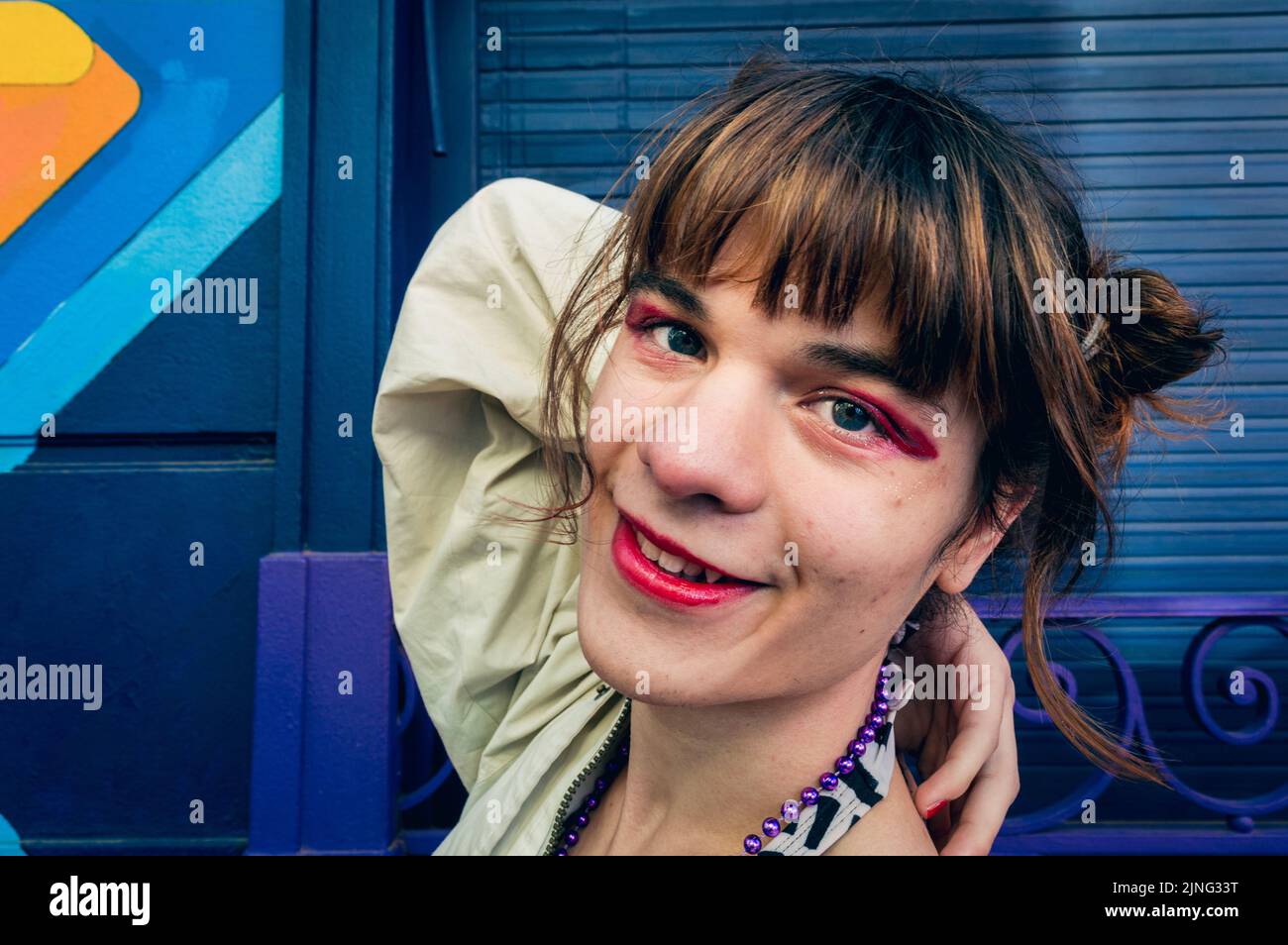 horizontal closeup portrait of happy transgender latin young woman with makeup looking at camera and smiling, posing standing in the street with blue Stock Photo