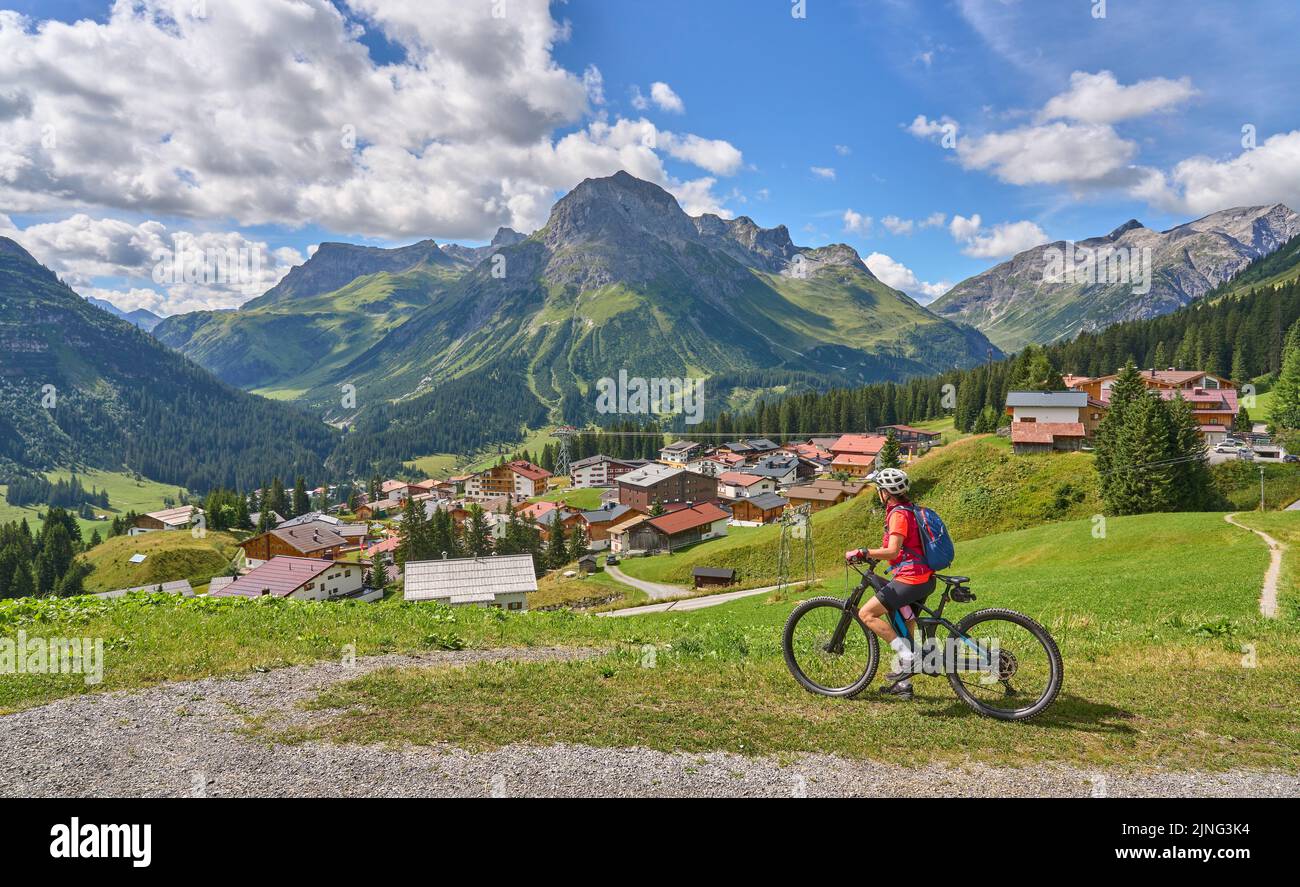 active senior woman, riding her electric mountain bike above famous village of Lech in the Arlberg area , Tirol, Austrian Alps Stock Photo