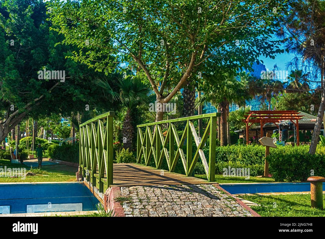 Beautiful summer landscape. View of the path and the bridge in a green garden with green trees along the embankment in Kemer, Turkey. In the backgroun Stock Photo