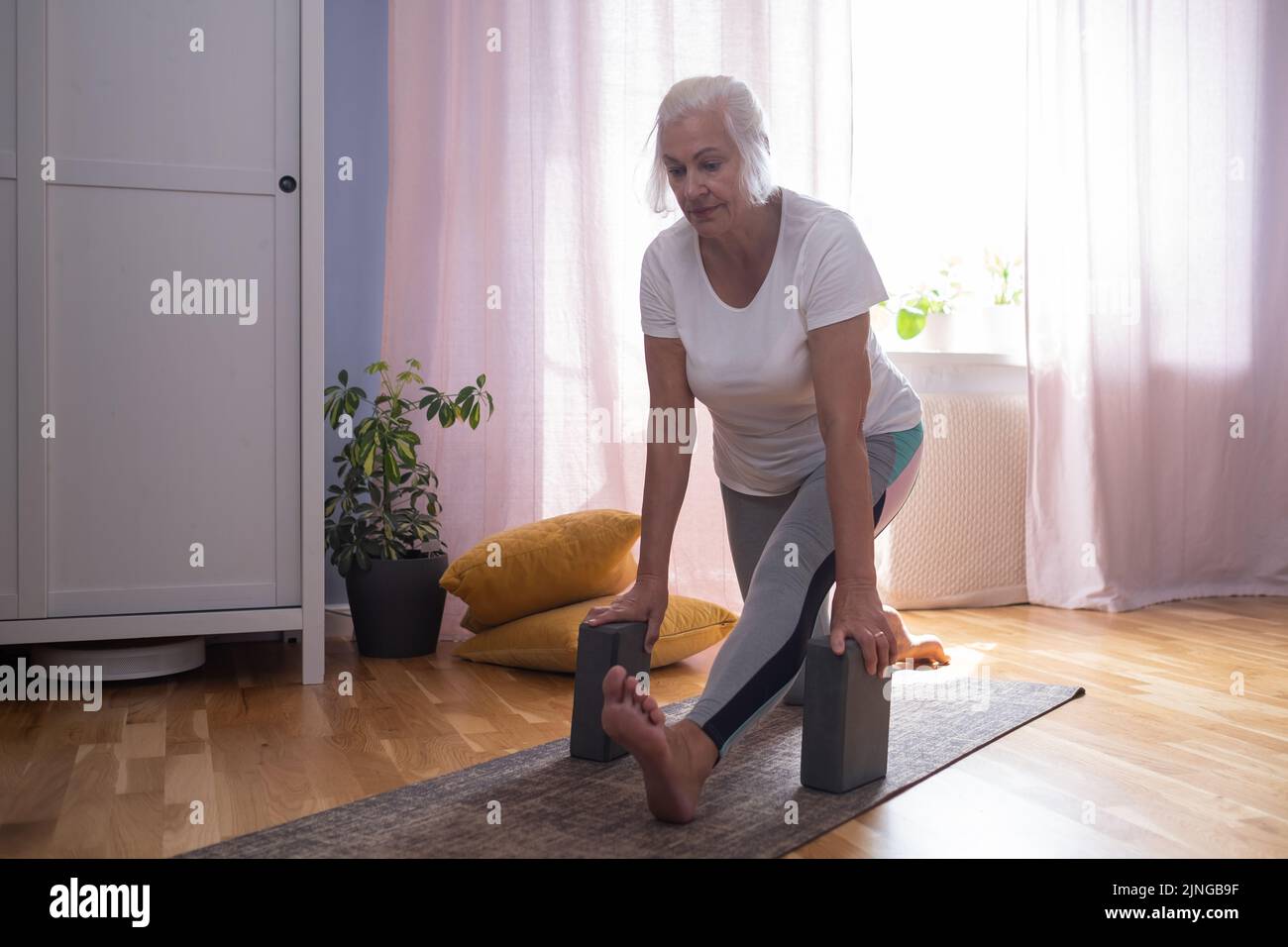 Senior aged woman sitting on the yoga mat and stretching her legs and back using blocks in the living room. Stock Photo