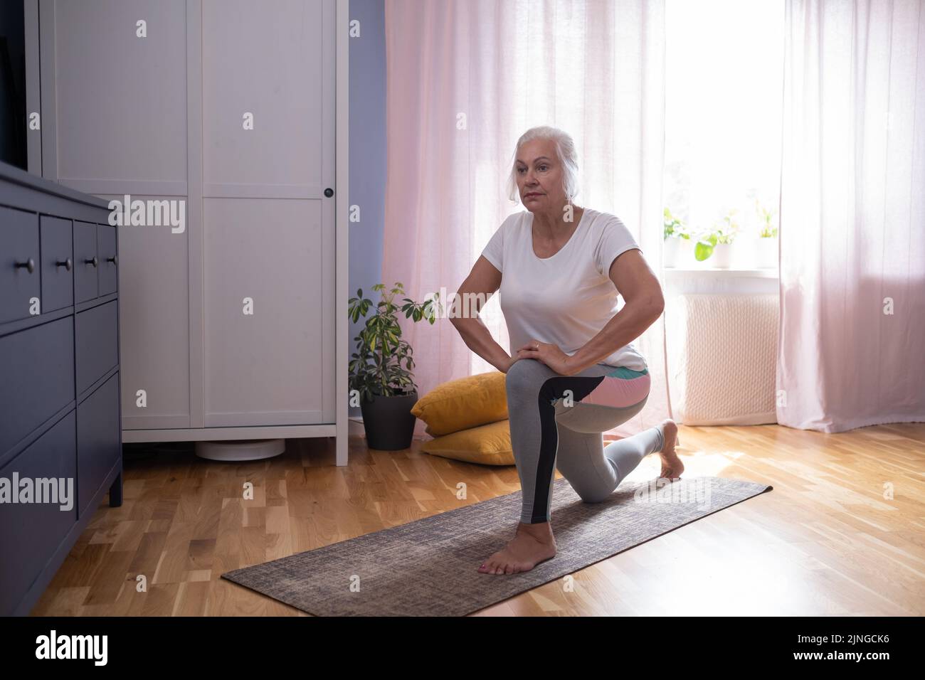 Senior aged woman sitting on the yoga mat and stretching her legs in the living room. Stock Photo