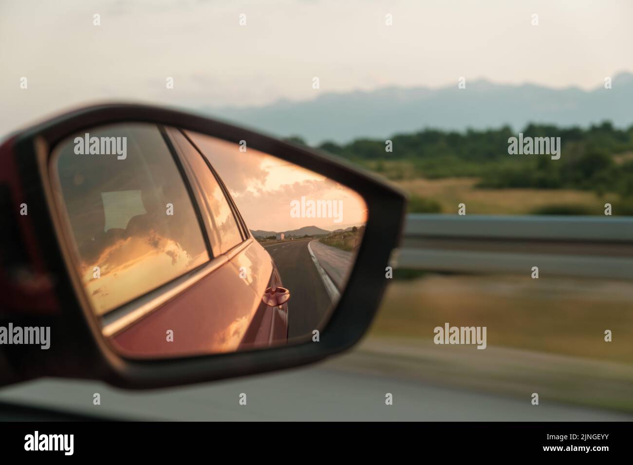 Red car drives past hilly landscape. Rear-view mirror shows roadway behind vehicle and door glass reflects sunset sky closeup Stock Photo