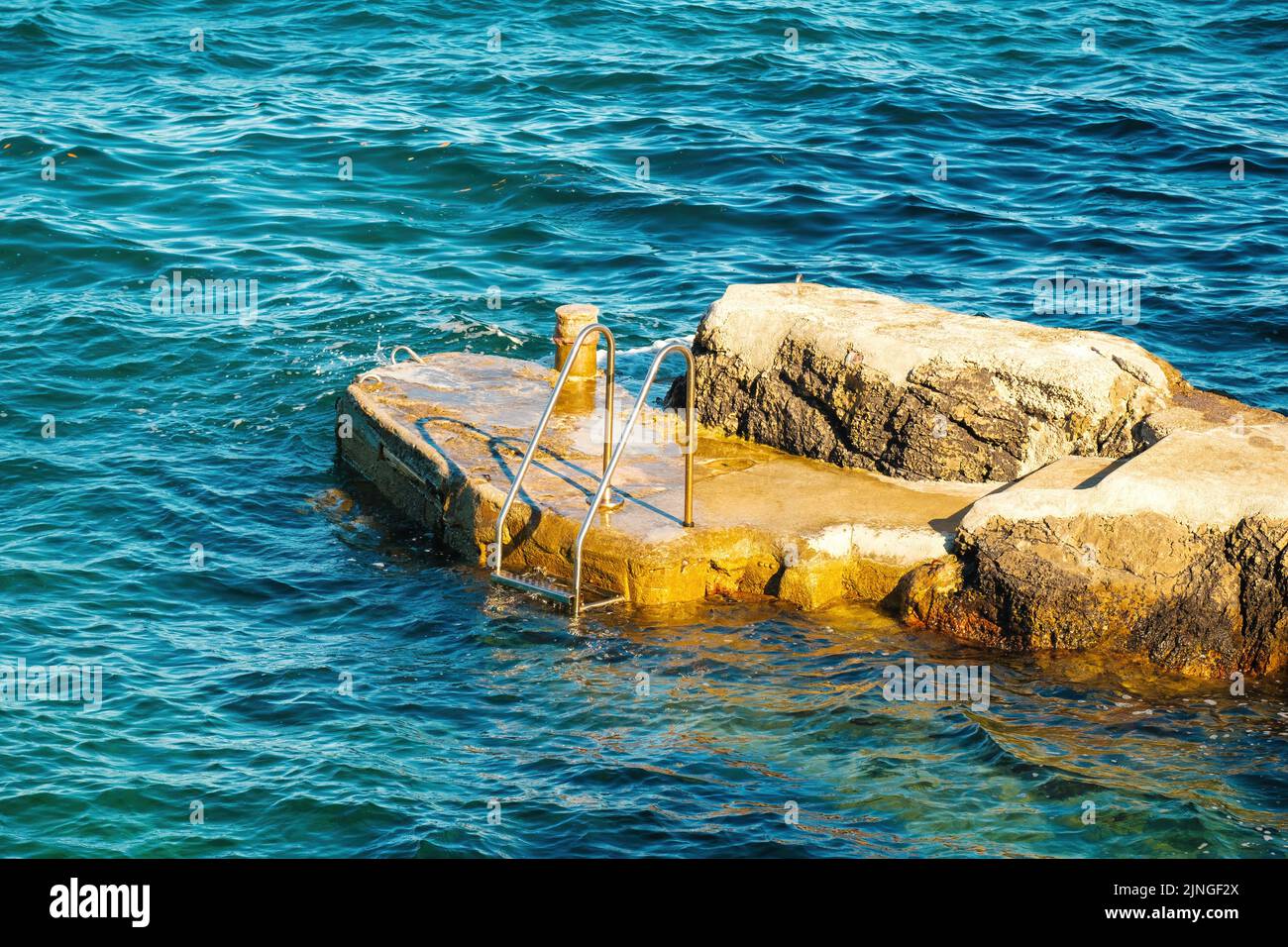 Metal ladder for swimmers descending into sea from small rock at sunlight. Stone pier washed by waves among deep turquoise sea at resort Stock Photo